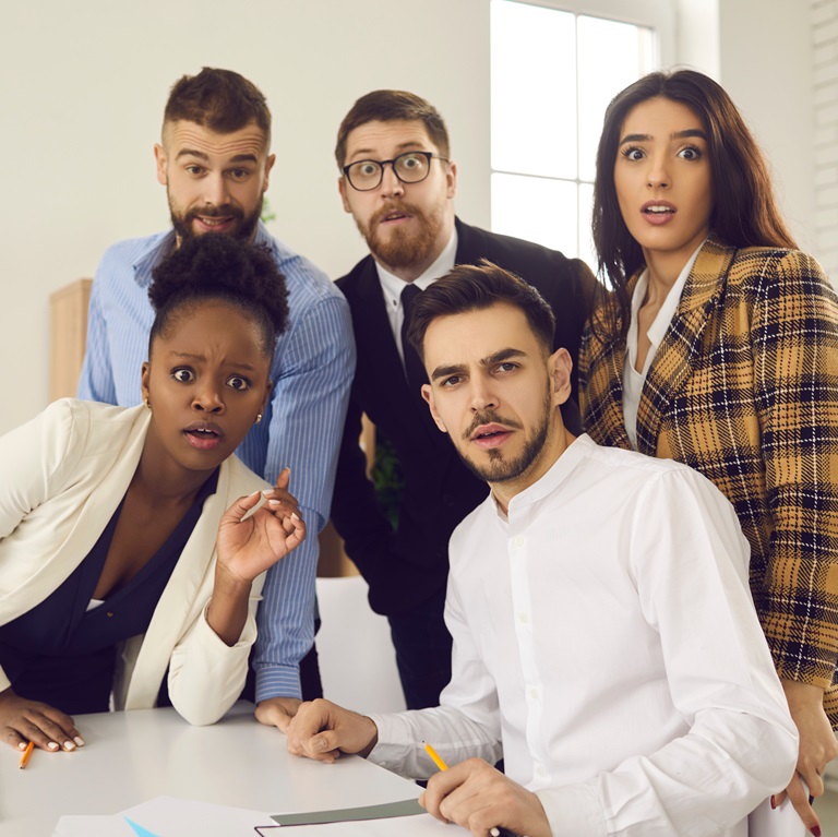 Five people in business attire gathered at a table stare at the camera with shocked faces after witnessing a business networking mistake.