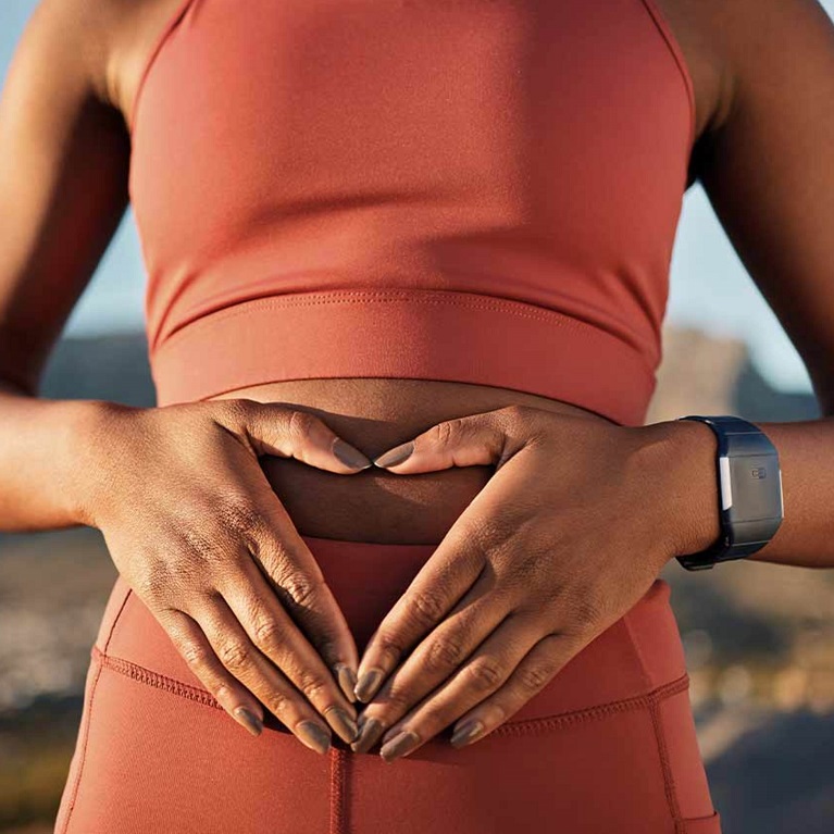 The midsection of a woman standing outside in workout clothes. Her hands are over her gut with tips of her fingers and her thumbs touching.