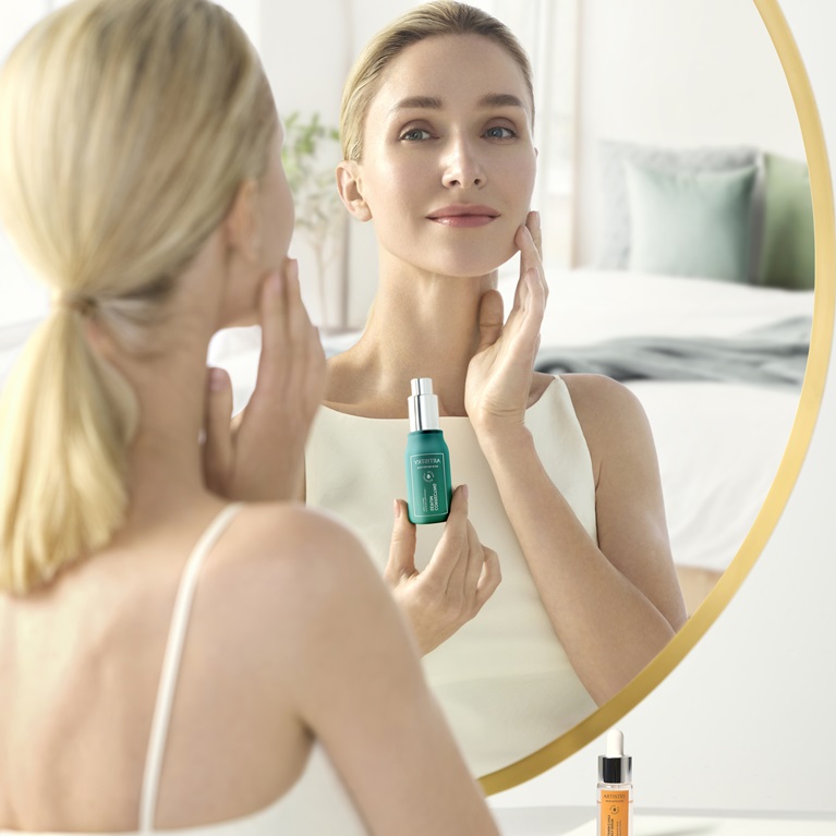 A woman looking in the mirror. She is holding a bottle of Artistry Skin Nutrition Correcting Serum in one hand and applying it to her cheek with the other hand.